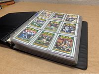 Opened black binder with 9 cards visible on one page, each card encased in plastic sleeve, showing front of cards with NFL players.