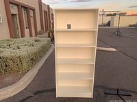Front view of a white bookshelf with five shelves standing outside.