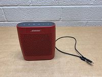 Front view of red Bose Bluetooth speaker with charging cable visible, showing front grille and logo.