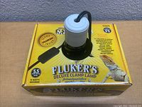 Fluker's Deluxe Clamp Lamp with ceramic socket, black clamp and switch on yellow branded box, showing overall condition and packaging.
