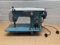 Front and side view of blue and white Kingston vintage sewing machine with power cord attached. Machine has knobs and dials, showing detailed controls for zig zag and stitch adjustment.