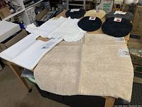 White bathroom rug with ruffled edges, beige large bath rug, and several oval toilet seat covers in black, beige, and gray colors laid out on table.