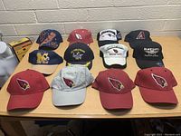 Twelve hats laid out on a table showing a mixture of Arizona Cardinals sports caps, Disney Mickey Mouse cap, USS Ronald Reagan hat, Texas Longhorns cap, NFL champions visors, and one 'Black Bart's Flagstaff Arizona' souvenir hat.
