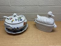 Two ceramic soup serving bowls on a table and against a wall: a white tureen with colorful floral design, lid, ladle, and matching plate alongside a white duck-shaped covered bowl.