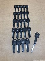 Photo shows 25 folding key cutters arranged on flat surface, all uniformly black plastic handles with folding blade keys extended on one item to reveal function.