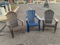 Three Adirondack style outdoor chairs in parking lot on asphalt. Two chairs are brown plastic, one chair is blue plastic. Chairs have slatted backs and wide armrests, typical style for Adirondack chairs.