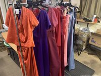 View of assorted formal dresses hanging on a rack showing various colors and styles including orange, purple, maroon, pink, and blue dresses.