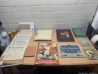 Photo of lot showing vintage newspapers, scrapbooks, hymn books, photo albums, and postcards arrayed on table.