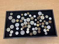 Tray with assorted old foreign coins in various sizes and metals spread out on black surface.