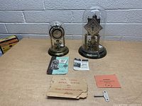 Pair of two vintage Schatz mid century clocks with glass domes and brass components, placed on a wooden surface along with multiple instruction manuals and a winding key.