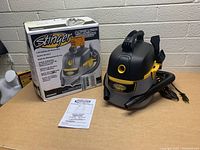 Shop vacuum, flexible hose, power cord, manual, and box displayed on table against cinderblock wall