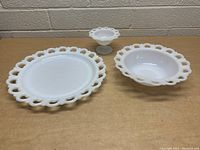 Photo showing three vintage milk glass items with open lace rims on a table: a large plate, a bowl, and a pedestal compote dish.
