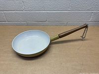 Side view of vintage Descoware frying pan with yellow exterior enamel and white interior, featuring a wooden handle with a metal chain.