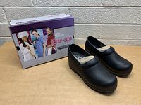 Pair of black Skechers Tone Up shoes with original box in background against white brick wall.