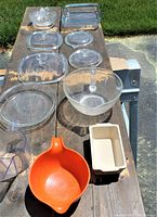 Top view of 11 kitchen items arranged on a bench outdoors, showing various glass lids, a glass juicer, a clear glass bowl, the orange plastic mixing bowl, and small ceramic dish.