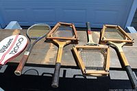 Four vintage tennis racquets laid out on a wooden table, including the Wilson T-2000 with its cover visible on the left. All racquets show varying wood tones and aged grips.