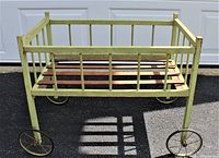 Lateral view of antique wooden doll bed showing yellow painted spindle rails, wooden slatted base, and four metal wheels.
