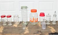 Full collection of 10 assorted vintage glass jars arranged in a row on wooden surface, visible lids and jar shapes
