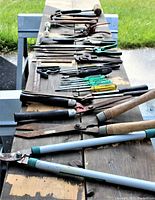 Photo showing assortment of 33 used hand tools laid out on wooden surface including screwdrivers, pliers, hammers, files, and hedge clippers, with some tools showing minor rust and wear.