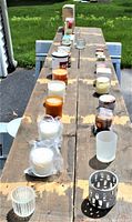 Long wooden table displaying 26 candles and votive holders arranged in two rows, showing various colors and holders