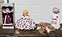 Overview of four dolls on a rustic wooden surface against a garage door backdrop showing boxed porcelain doll, vinyl doll, nurse mouse doll, and country angel decoration.