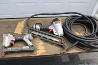 Two Porter-Cable pneumatic nailers side by side on a wooden surface, one the NS100B crown stapler, the other DA250C finish nailer, plus a coiled black air hose.