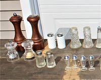 Photo showing two wooden salt and pepper mills, assorted glass salt and pepper shakers including clear glass, white textured, and vintage style with metal tops.