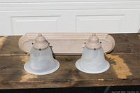 Front view of the vanity wall sconce light showing two frosted bell-shaped glass shades mounted on beige wooden mounts and a decorative wooden backplate.
