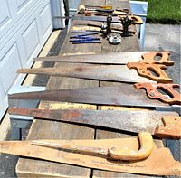 Photo showing vintage saws with wooden handles, rusty blades, and various hand tools arranged on a wooden surface outdoors.