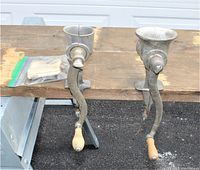 Two vintage crank meat grinders side by side on a wooden surface with handles visible, showing their metal bodies and wooden crank handles.