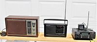 Front view of three vintage radios: Realistic wooden AM/FM radio, Emerson black weather/AM/FM/TV radio with telescopic antenna, RadioShack Pro-2050 scanner with antenna and power cord