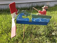 Side view of wooden whirlygig showing fisherman in boat with fish and red/white propeller on wooden stake in grass.