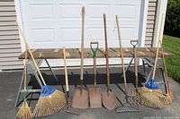 Photo showing four bamboo leaf rakes with blue plastic tine heads, one metal bow rake, three shovels with different head shapes, one pitchfork, and one wooden scythe.