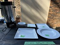 Photo showing large stainless steel coffee urn with black base and spout next to white square and round plates and one bowl.