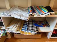 Overview of multiple folded fabric pieces including leaf pattern, stripes, and checkered designs on white shelving unit.