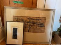 Two framed artworks leaning against wall; one large silver-framed sepia landscape and one smaller black-framed vertical blue print