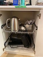 Full view inside kitchen cabinet showing all small appliances including Oster kettle, PC coffee grinder, Bron hand mixer with accessories, Breville Panini maker, and DeLonghi grill below