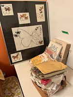 Framed butterfly needlework artwork and pile of assorted needlework supplies, books, and fabric on a table