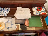 Top drawer contents showing folded tablecloths, embroidered white napkins, patterned cloths, and placemats in various colors.