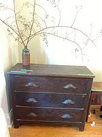 Full view of dark wooden dresser with three drawers and metal handles, ceramic vase with dried branches on top.