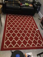 Full view of the red and beige area rug on the floor beside a black leather couch and side table.