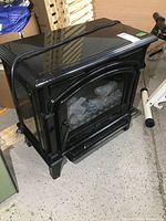 Front and side views of a black electric freestanding fireplace with glass front showing realistic artificial logs inside, placed on a concrete floor.