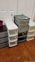 Overall view of four different sized plastic tote drawer units in white, black, and grey colors with small red metal bucket on top of black drawer unit.