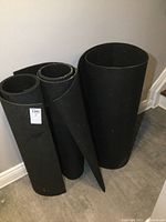 Photo showing all three large rolled black rubber mats standing against a wall on a tiled floor.