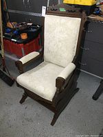 Front angled view of antique Eastlake style spring rocker showing carved wooden frame, padded seat, backrest, and armrests with cream floral patterned upholstery.