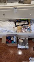 Overall view of drawer with white linens, framed quote, collectors plate, boxed candle, and a box of decorative balls on floor.