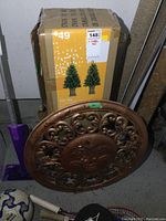 Metal sun face outdoor art piece showing face and scrollwork with some paint chipping, placed on floor near boxed Christmas trees and sports gear.