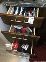 Three kitchen drawers open showing various cutlery, utensils, kitchen tools, red oven mitt, and towels in good to worn condition