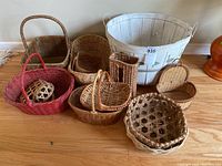 Photo showing eleven wicker baskets in various shapes and colors (natural and red), one large white painted bucket with wire handles and a number tag, all sitting on a hardwood floor.