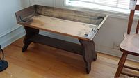 Front view of an antique wooden bench with a darker stained base and light natural wood seat showing wear and stains.